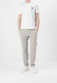 Hombre vistiendo una camiseta de manga corta blanca con un diseño de frutas, pantalones gris claro con las manos en los bolsillos y zapatillas blancas, de pie frente a un fondo blanco.