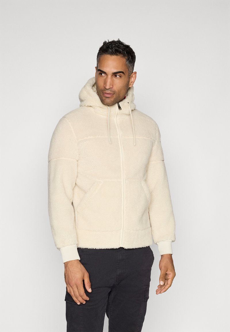 Mann trägt eine cremefarbene Fleecejacke mit Reißverschluss und vorderen Taschen, steht und schaut zur Seite, vor einem schlichten Hintergrund.