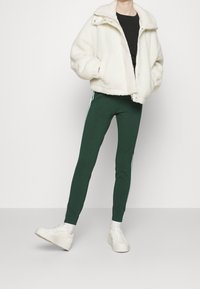 Vit fuskpälsjacka med hög krage, kombinerad med mörkgröna leggings med vita sidostråkar och vita sneakers.