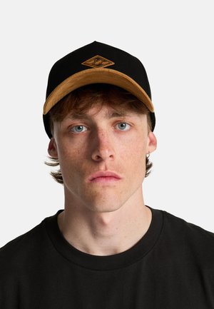 Jonge man met een lichte huid en sproeten draagt een zwart-beige honkbalpet en een zwart shirt, kijkt recht vooruit met een neutrale gezichtsuitdrukking.