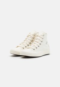 Zapatillas altas de lona color beige claro con ojales plateados, punta redonda, suela de goma plana y detalles bordados en el lado exterior.
