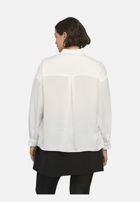 Blusa bianca a maniche lunghe in un tessuto leggero, con un collo modesto e un dettaglio plissettato sul retro, abbinata a una gonna mini scura.