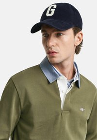 Olive green polo with a denim-blue collar and white placket, featuring a small logo on the chest and a navy corduroy cap with a letter 'G.'