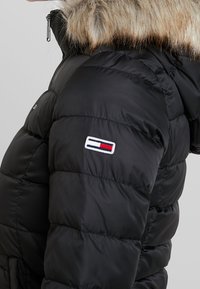 Czarna kurtka puchowa z błyszczącym, pikowanym wykończeniem i kapturem z sztucznym futrem. Posiada haftowane logo w czerwonym, białym i niebieskim kolorze na rękawie.