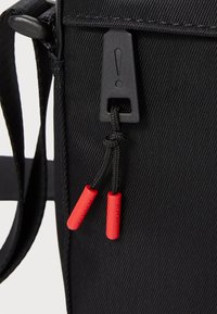 Čierna látková taška so zipsom, ktorá má čierny úchyt označený výkričníkom a červené konce šnúrky.