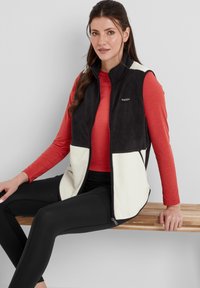Black and cream sleeveless vest with front zipper, contrasting fleece texture, worn over a red long-sleeve shirt and black leggings.