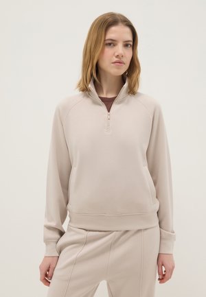 Junge Frau mit schulterlangem blondem Haar, die einen beigen Sweatshirt mit Viertel-Reißverschluss und passende Hosen trägt und vor einem schlichten Hintergrund steht.