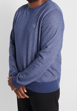 Homme portant un sweat-shirt bleu à col rond avec un petit logo sur la poitrine, debout, les mains jointes devant, sur un fond uni.