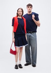 Γυναίκα και άνδρας στέκονται δίπλα δίπλα φορώντας μπλε σκούρα μπλουζάκια, η γυναίκα με κόκκινο πουλόβερ περασμένο στους ώμους και τσάντα, ο άνδρας κρατάει μπλε καρό πουκάμισο περασμένο στον ώμο.