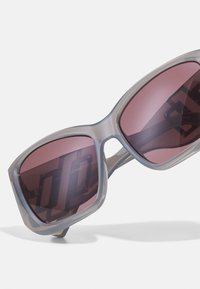 Sivé oversized slnečné okuliare s tmavo ružovými sklami, s transparentným rámom a viditeľnými prvkami hardvéru. Moderný geometrický dizajn.