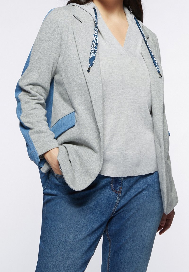 Tenue décontractée avec un sweat à capuche gris clair muni de cordons bleus, un blazer gris assorti avec des accents bleus, et un jean bleu.