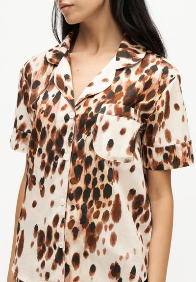 Femme portant une chemise à manches courtes boutonnée avec un imprimé animal marron, noir et beige et une poche poitrine.