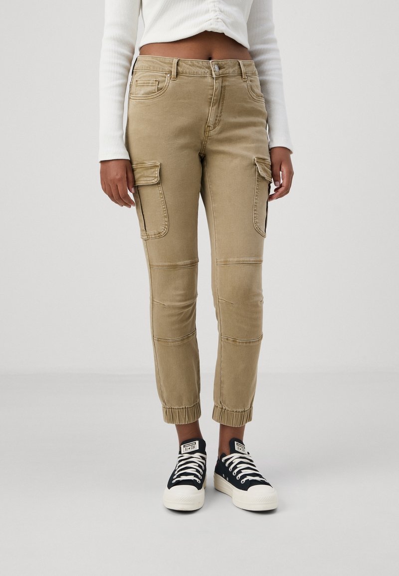 Femme portant un pantalon cargo beige avec des poignets élastiques, associé à des baskets basses noires et blanches, debout devant un fond uni.