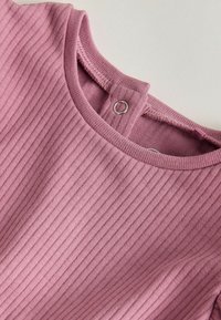 Lahka tkanina vijolične barve z rebrasto teksturo, okrolim izrezom in zapiranjem na pritrditev z okrasnim kovinskim obročkom, ki poudarja teksturiran zaključek.