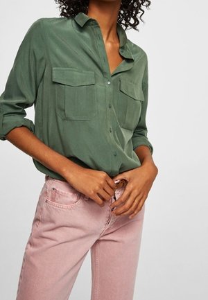 Kvinde iført en grøn skjorte med knapper, brystlommer og opkrogede ærmer, der spænder et par lyserøde, højttaljede jeans med bælte.