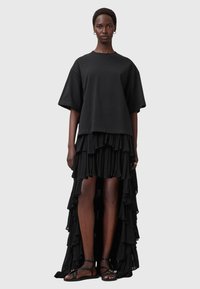 Sort kjole, der kombinerer en kort, oversized top med en draperet ruffleskirt. Har et lagdelt design og åbne sider. Flade sorte sandaler.