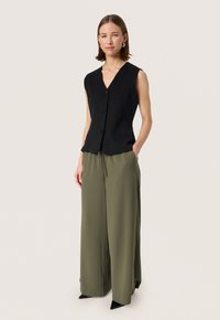 Black sleeveless vest with a V-neck, featuring front buttons. Paired with loose olive green trousers and pointed black shoes.
