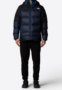 Veste matelassée bleue et noire avec capuche, présentant des sections matelassées et une fermeture éclair frontale. Portée avec un pantalon noir et des baskets foncées.