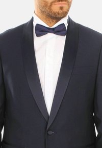 Giacca da smoking di colore navy con rever in satin, tessuto a trama, chiusura a un bottone, abbinata a una camicia bianca e cravatta a farfalla scura.