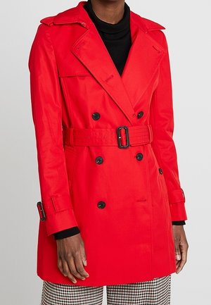 Femme portant un trench-coat rouge vif à double boutonnage avec des boutons noirs et une ceinture à la taille, par-dessus un haut noir et un pantalon à carreaux.