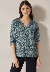 Blusa di colore teal scuro con motivo floreale, scollo a V e maniche arrotolate, realizzata in un tessuto leggero, con una vestibilità rilassata e un leggero lucido.