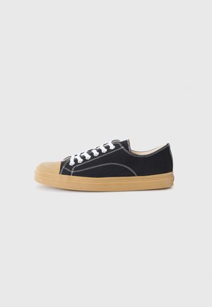 Svarta kanvas sneakers med vita snören, en rund beige gummitå, och en gummisul. Vita sömmar framhäver designen.