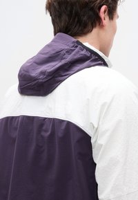 Νεαρός άνδρας με λευκό και μωβ μπουφάν North Face με κουκούλα, εμφανιζόμενος από πίσω και ελαφρώς στραμμένος προς τα δεξιά.