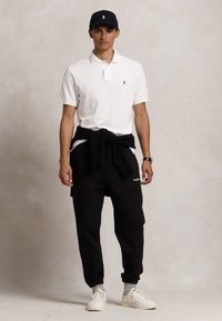 Weißes Poloshirt, schwarze Sweatpants mit Logo, schwarzer Sweater um die Taille gebunden, weiße Sneaker und eine schwarze Mütze. Neutraler Hintergrund.