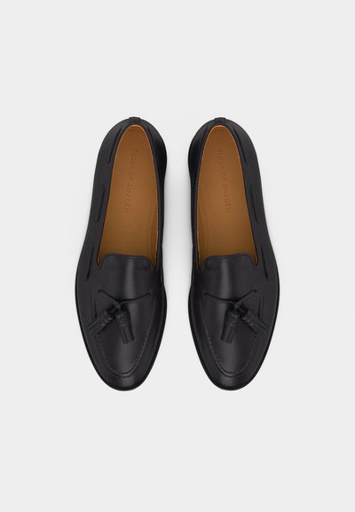 Fekete bőr mokaszin pár bojttal és barna talpbetéttel, felülről fehér háttéren nézve.