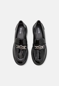 Mocassins en cuir verni noir avec un bout arrondi, un accent en chaîne argentée et un détail de logo sur la bride. Texture lisse avec une finition brillante.