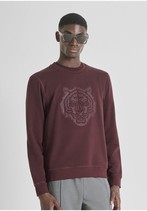 Bordo dukserica s teksturiranim grafikom tigra na prednjoj strani. Dugi rukavi, okrugli izrez i opušten kroj. Uparena s sivim hlačama.
