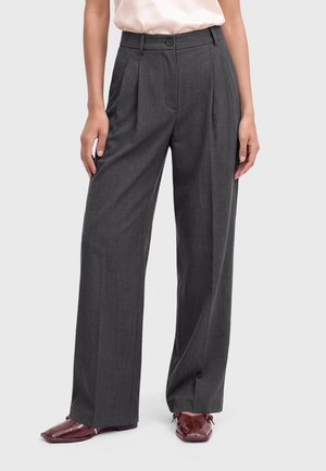 Femme portant un pantalon taille haute gris foncé à jambes larges et des chaussures bordeaux à bout pointu, debout devant un fond uni.
