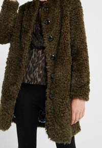 Olivgrüner Kunstpelzmantel mit strukturiertem, flauschigem Material. Verfügt über schwarze Knöpfe und eine leicht übergroße Passform. Innenfutter mit blauem Muster.