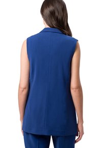 Femme aux longs cheveux bruns portant un blazer bleu sans manches et un pantalon bleu assorti, vue de dos sur un fond blanc.