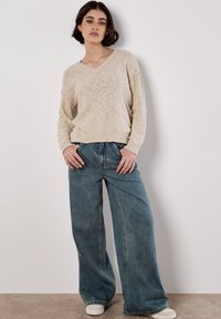 Beige gebreide v-hals trui met verlaagde schouders, gecombineerd met een losse spijkerbroek in blauw en witte sneakers, geplaatst tegen een neutrale achtergrond.