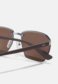 Ochelari de soare dreptunghiulari, căprui, cu o ramă din metal argintiu și lentile căprui. Au sprijină gri cu un accent de logo. Include pernuțe de nas transparente.