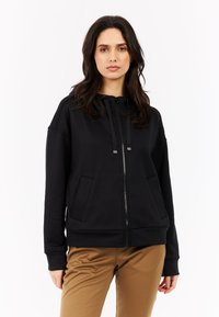 Zwarte zip-up sweatshirt met kangoeroe zakken, geribbelde manchetten en een trekkoord capuchon. Gemaakt van gladde stof met een casual pasvorm, gecombineerd met beige broek.