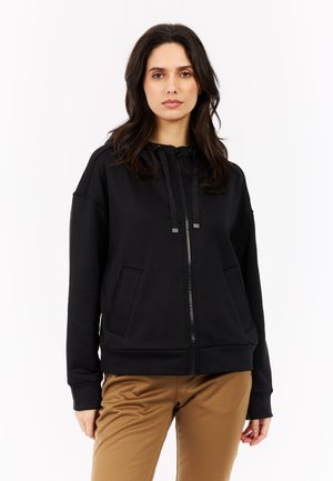 Zwarte zip-up sweatshirt met kangoeroe zakken, geribbelde manchetten en een trekkoord capuchon. Gemaakt van gladde stof met een casual pasvorm, gecombineerd met beige broek.