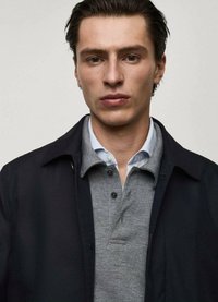 Svart ytterjacka över en grå pikétröja med krage och två knappar. Tröjan är lager-på-lager över en ljusblå randig skjorta.