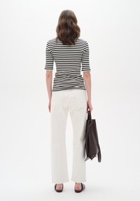 Frau steht mit dem Rücken zu uns in einem schwarz-weißen gestreiften Top, weißen Hosen und schwarzen Schuhen, hält eine große dunkelbraune Handtasche vor einem weißen Hintergrund.