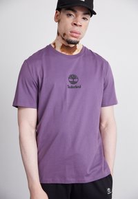 Violetinis medvilninis marškinėliai su trumpomis rankovėmis, ant krūtinės puikuojasi juodas Timberland logotipas, derinami su juodomis kelnėmis.