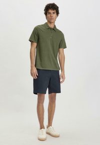 Olijfgroen polo shirt met korte mouwen en een kraag, gecombineerd met marineblauwe shorts. Model draagt witte sneakers. Eenvoudig, strak ontwerp.