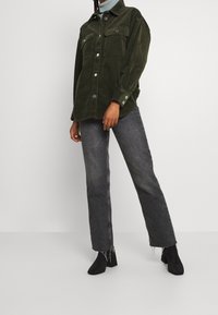 Zelená košile z manšestru s dvěma kapsami na prsou, doplněná vybledlými šedými džíny s rovnými nohavicemi a černými kotníkovými botami.