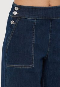 Pantalones de mezclilla oscuros de tiro alto que presentan tres botones plateados en la cintura, con costuras amarillas contrastantes y un bolsillo lateral.