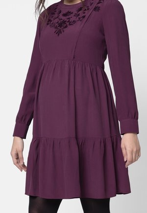 Robe prune à manches longues avec volants froncés et broderies florales sur la poitrine, portée avec des collants noirs sur un fond neutre.