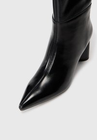 Bota negra de cuero hasta el tobillo con punta afilada y tacón cuadrado. Presenta una textura suave y costuras mínimas a lo largo del lado.