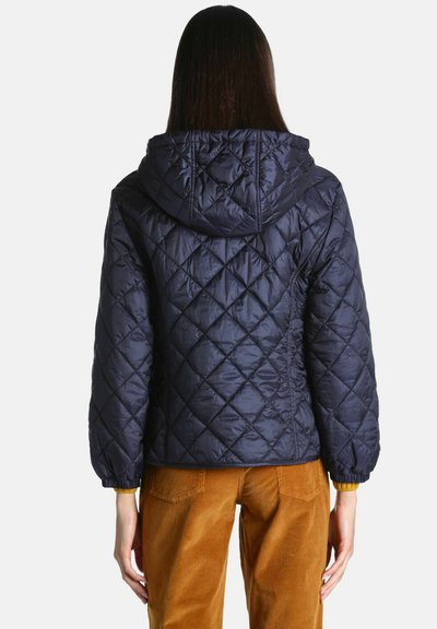 Personne aux longs cheveux foncés portant une veste matelassée à capuche bleu marine et un pantalon couleur moutarde, vue de dos sur un fond uni.
