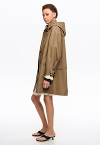 Young person in brown hooded raincoat, white lace-trimmed dress, and black open-toe kitten-heel sandals standing sideways on white background.