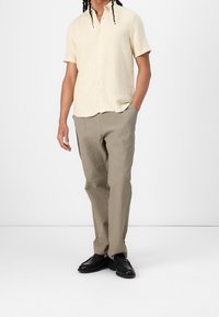 Kortærmet cremet skjorte med knaplukket krave, kombineret med teksturerede lysebrune bukser. Sorte sko fuldender outfittet.