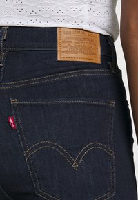 Tmavě modré džíny z denimu s koženou nášivkou na zadní straně, která obsahuje embosovaný text. Viditelné žluté prošití a charakteristický zkroucený design kapes.